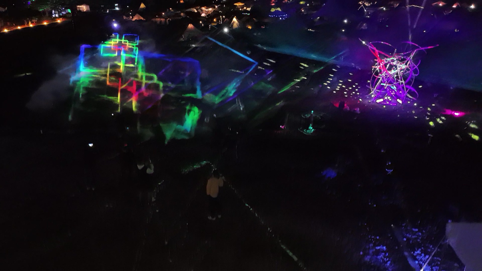 A night drone view over the Re:birth Festival grounds in Japan: coloured light lattices and a glowing lattice sculpture among the festival lights