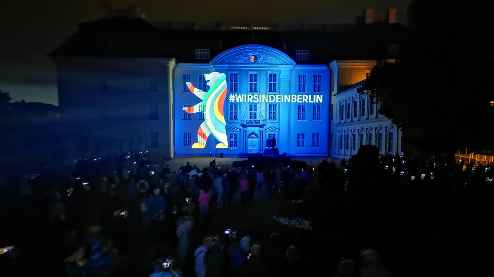 Schloss Köpenick in Berlin at night during the Festival of Lights 2020, its façade lit blue by a projection of the Berlin bear and #WirSindEinBerlin, with the audience in front