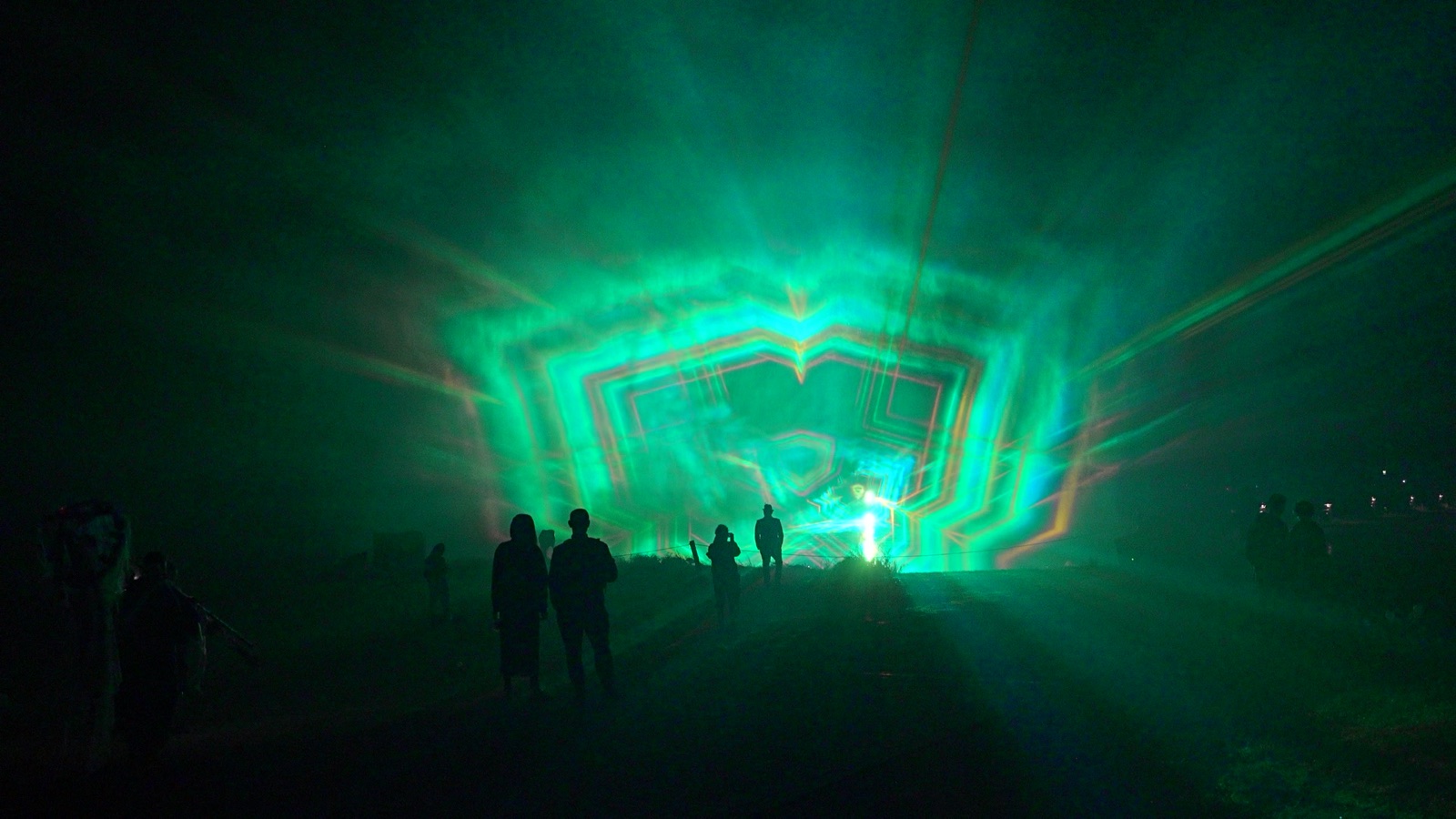 Beams of blue and orange light passing through layered water and mist screens at night, with festival visitors silhouetted in front of the Depth/100 installation at Ozora