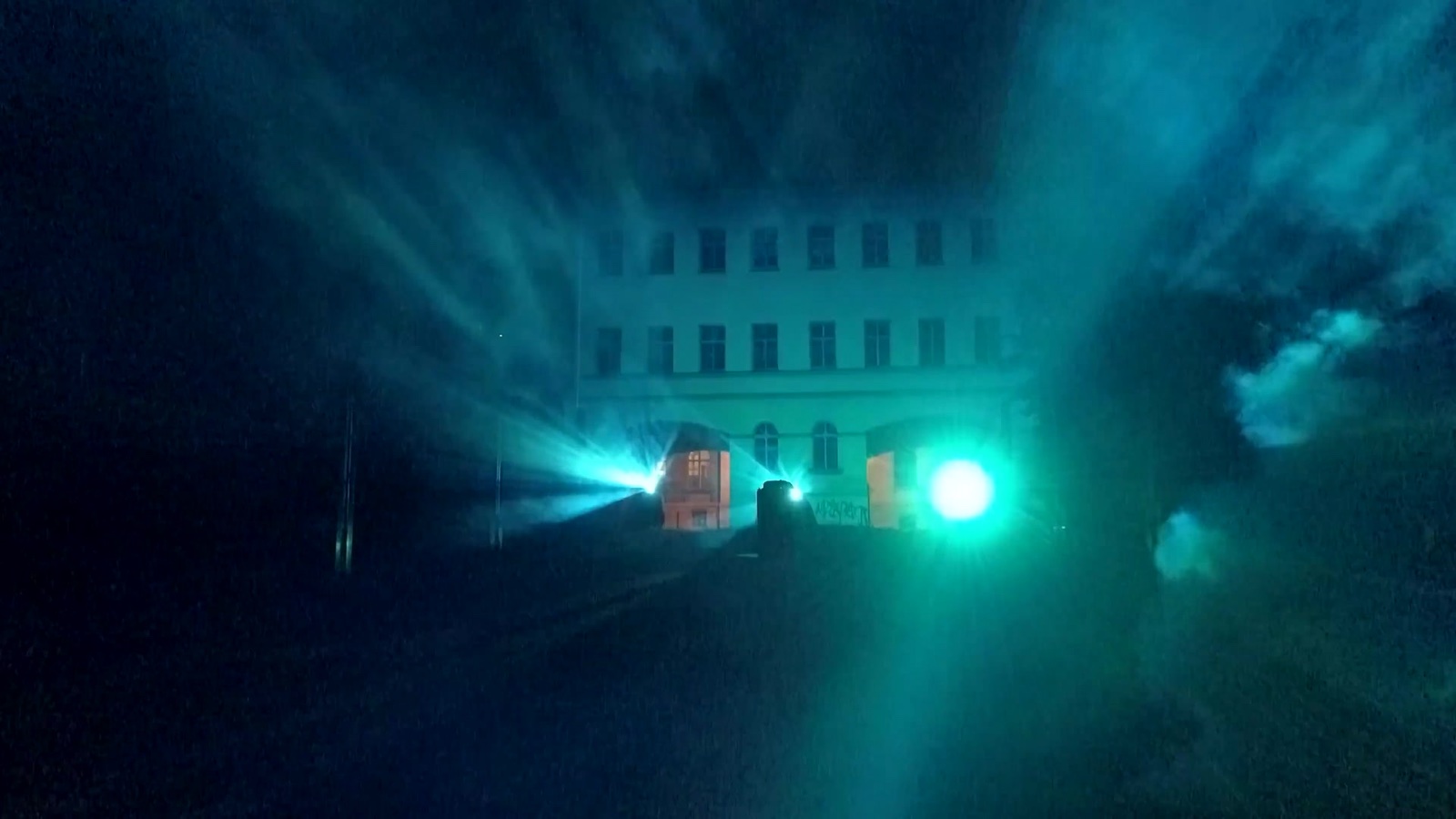A crowd at night in front of a mapped building at Boom Festival, with beams of blue and white light passing through fog in front of the façade